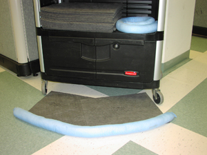 A cylindrical tube of absorbent batting attached to absorbent floor matting is on the ground in front of a wheeled cart.  Additional materials of the same sort are stored on the cart.