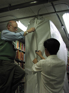 Two men are securing plastic sheeting with binding clips around affected shelving areas.  One is on a ladder securing the top of the sheeting, while the other is assisting from ground level.