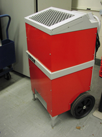 A red and grey dehumidifier is sitting on the floor in the disaster cage.  The dehumidifier has two wheels on the bottom, and is meant to stand upright.  The top has a wire grate.  The electrical cord is wound neatly at the back near the handle.