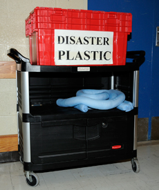 A bright red plastic bin is clearly labeled 'Disaster Plastic.' It is sitting on the top of a movable cart, ready for transport in case of an emergency. In the middle of the cart is absorbent tubing.