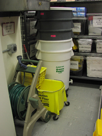 Five large plastic trash cans are stacked in a corner of the disaster cage.  In front of the trash cans are a mop bucket and a garden hose on a mobile reel.  A clearly labeled 220 volt outlet is on the wall above the garden hose.  Additional supplies, also labeled, are stored on metal shelving in behind the trash cans.