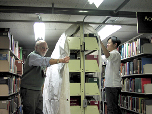 Two men are hanging plastic over a shelf of books.  One man is standing on each side of the shelf.  They are arranging the plastic so that it is centered on the top of the shelf and hangs down to the floor on either side of the shelf.  Extra plastic has been left so that the surplus can cover the end of the shelf.