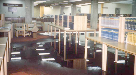 Color photograph of the National Library of Medicine's main reading room featuring several wooden study carrels with reference materials on shelves. A large portion of the floor is covered in water. The chairs have been moved behind absorbent tubing on the floor used to wick up the water.