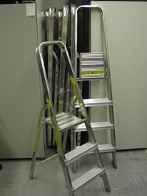 This photo has two metal step ladders, one with two steps and one with three.  One is set up and the other is leaning against a wall.  Three additional ladders are folded up and stored in a nook in the wall.