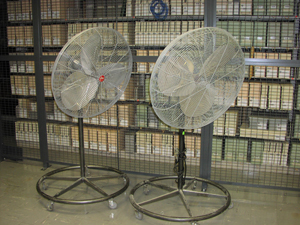Free standing floor fans are sitting in a collection area.  The fans' bases are on wheels for ease of movement in stack areas.  The cords are secured off of the floor