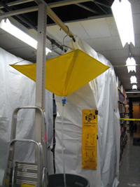 Picture of a leak diverter hanging from a sturdy ceiling support.  The plastic tube extending from the bottom is resting in a waterproof container.  Nearby shelving is covered in plastic sheeting.