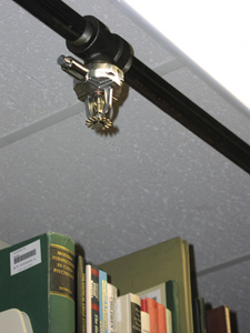 Photo of a sprinkler head hanging from the ceiling.  The sprinkler head protrudes from a pipe mounted parallel to the ceiling.  It is located over a shelf of books.