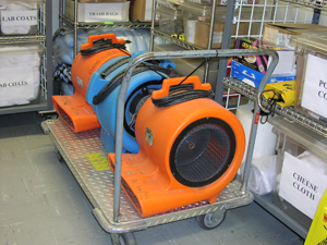 Three Squirrel Cage Fans are sitting on a rolling utility cart. The cords of the fans are secured and the fans themselves are attached to the handles of the cart with a bungee cord.  Surrounding the cart on two sides are wire shelving units with additional disaster supplies.  These are labeled with signs that say 'Lab Coats' 'Trash Bags' and 'Cheese Cloth'.