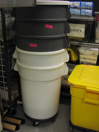 Five trash cans are stacked in the corner of a disaster cage.  Each trash can has wheels attached to the bottom of it for ease of movement. Next to the cans is a plastic bucket, and behind them is metal shelving with additional disaster supplies.