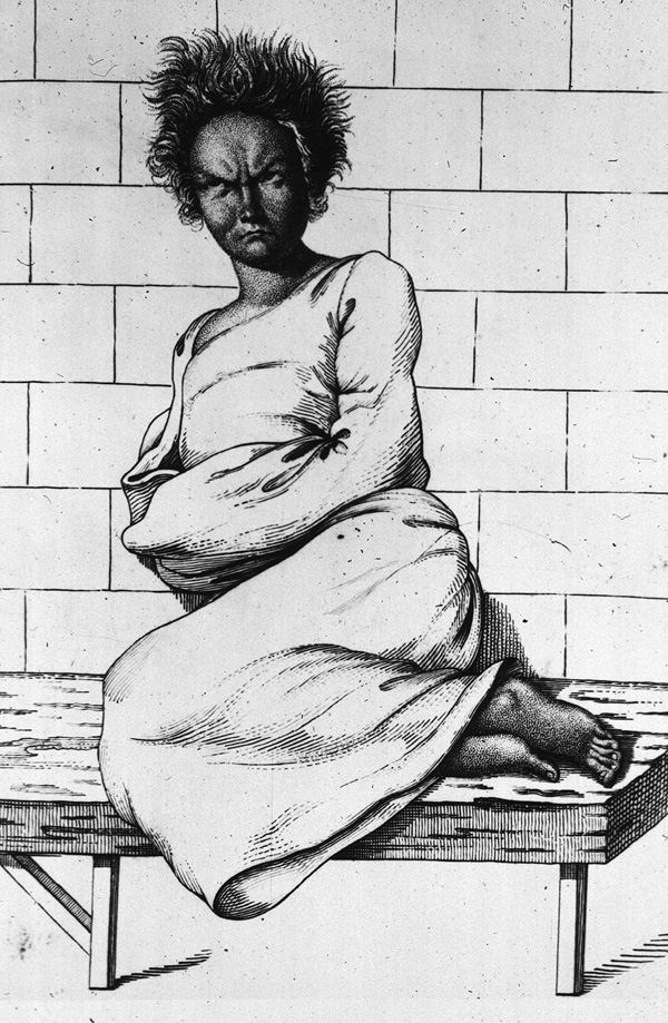 A scowling woman with her arms held in a gown sitting with her feet drawn up on a bench, and leaning against a tiled wall.