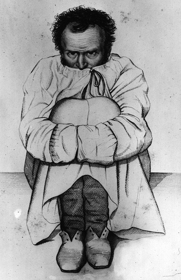 A man sitting on the floor hugging his knees wearing a robe, his hands tucked in the sleeves and the collar up over his mouth.