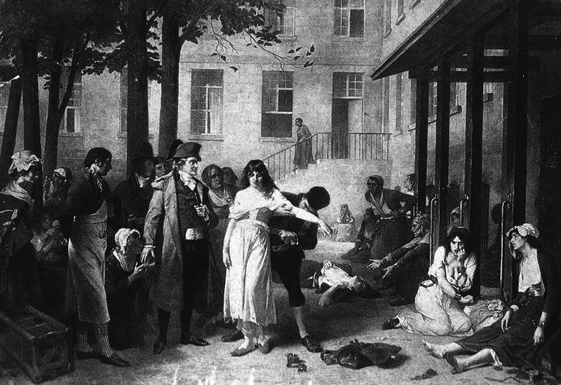 A man stands in the courtyard as a woman patient is freed from her chains; other women patients are chained to wooden posts attached to a building.