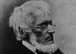 Formal photographic portrait an older, bearded, white man, seated, wearing glasses.