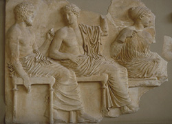Photograph of a frieze of the Parthenon showing Apollo, seated, wearing a toga and facing left.