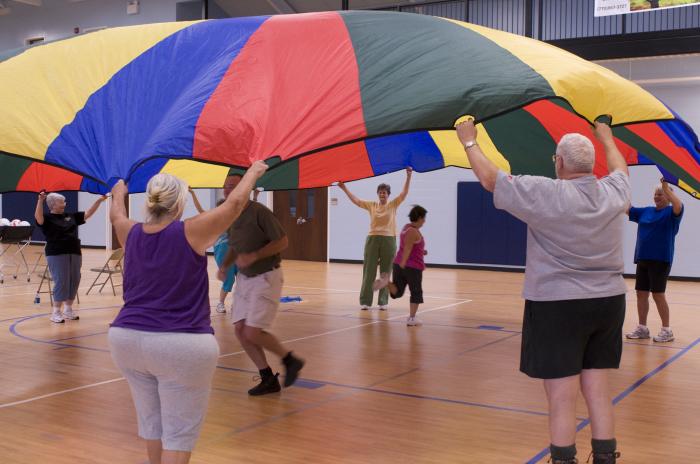 Senior men and women are in a gymnasium doing an activity where their arms are lifted up as they hold onto a parachute, while one man and one woman are running under the parachute.