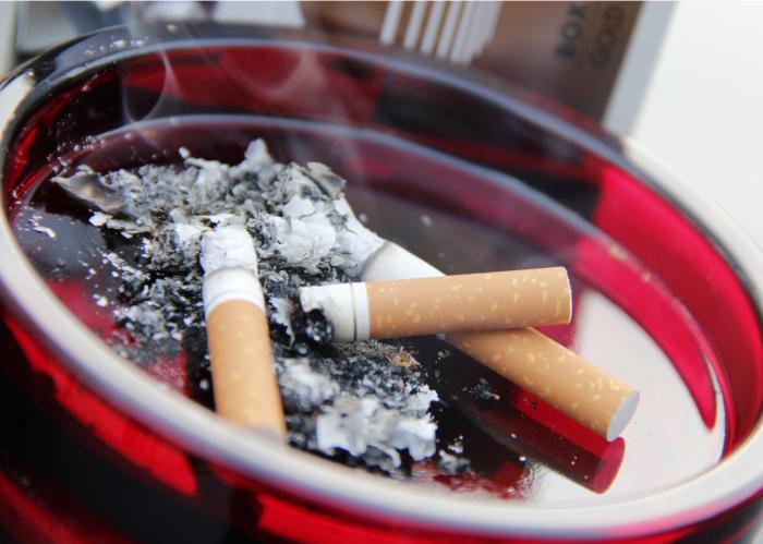 Three cigarettes are left burning in a round red ashtray, with smoke rising from the cigarettes. The cigarettes are almost completely burned down to the ends, and ashes are left behind in the ashtray.