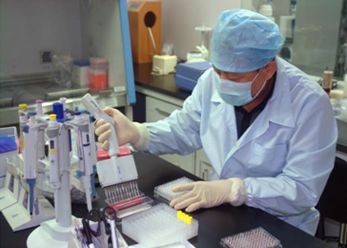 A medical lab technician, wearing a white coat, a blue paper surgical hat and mask, and protective gloves, is working in a lab.