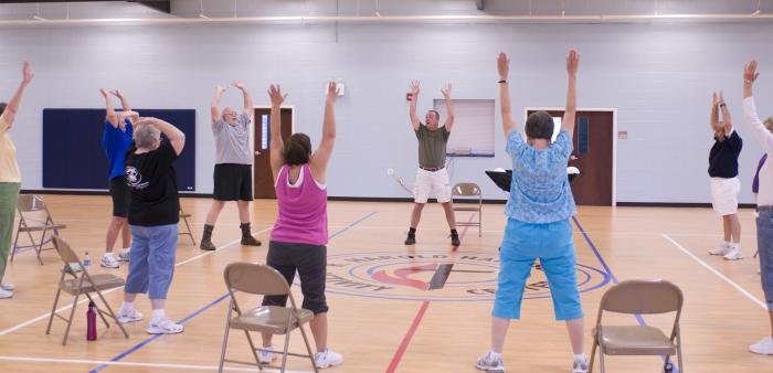 A senior fitness class taking place in a gymnasium with the participants in a circle, each standing in front of a folding chair, stretching with arms lifted up high