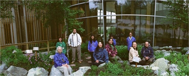 Color image of a group of individuals outside in a garden. Some are standing while others are sitting.