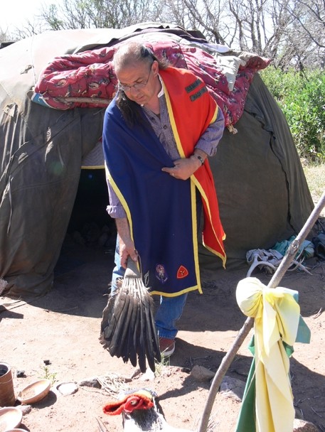 Color image of a man standing in front of a modern-day sweatlodge, bending over with a fan made of eagle feathers in his right hand.