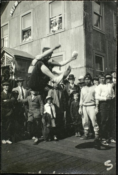 Black and white photograph of a male in mid-air performing a high kick and surrounded by many onlookers.