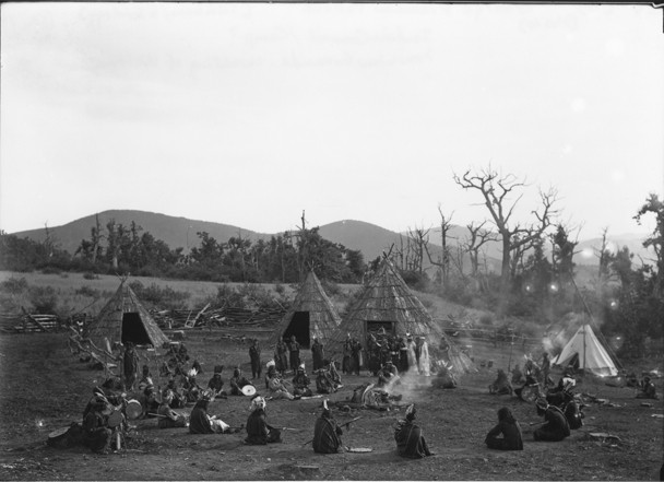 Black and white photograph of several Native Americans sitting down in a circle around a fire with four erected teepees in the background.