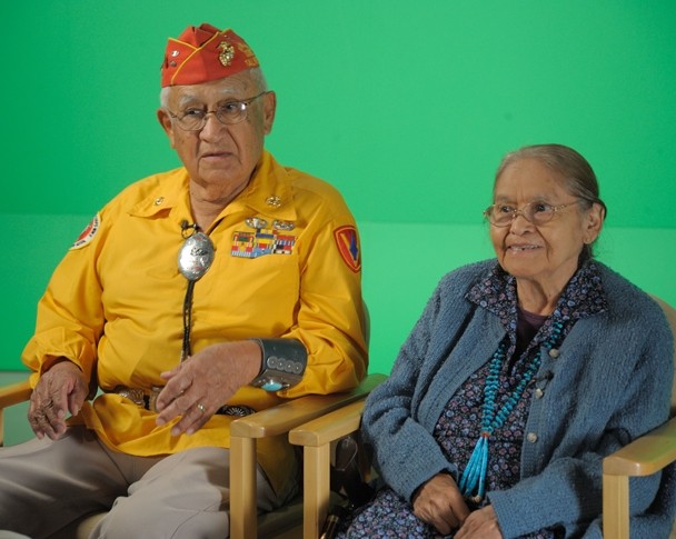 Color photograph of an elderly man and woman sitting next to each other in separate chairs in front of a green screen.