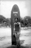 Black and white photograph of Duke Kahanamoku standing in front of his surfboard, which is inscribed with the word “Duke.”