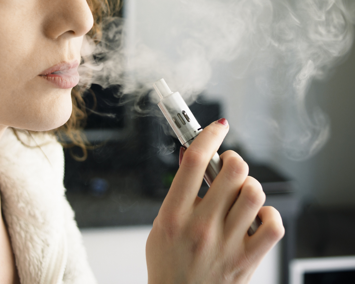 Close-up of a young woman exhaling smoke vapour from an electronic cigarette, holding the device in her hand. Shot at home, indoors.